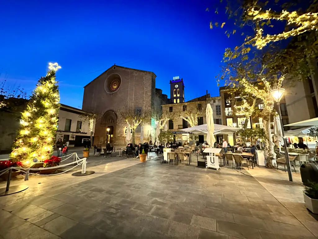 Pollença's Plaça Major at dusk with a lit Christmas tree in front of the church